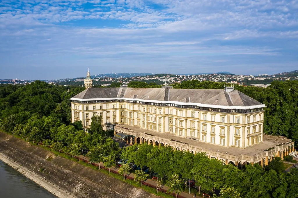 Légi felvétel az Ensana Grand Margitszigetről, ahol a patinás krémszínű épület látható a Duna partján, erdők és Budapest panorámájával